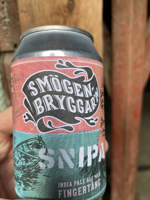 En ölburk med etiketten "Smögen Bryggar'n" och texten "SNIPA India Pale Ale med Fingertång" hålls i en hand mot en träbakgrund.