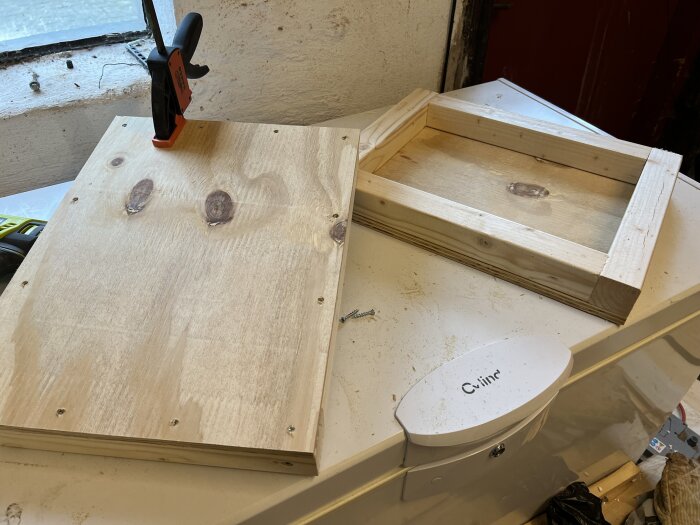 Två träramar av 45x45-reglar ligger på en arbetsbänk, varav en är hopsatt med plywood och en tving.