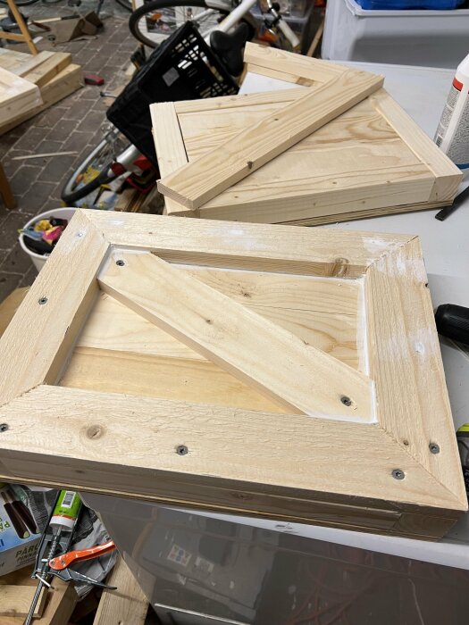 Två träramar med diagonalstöttor under tillverkning, stående på ett arbetsbord med verktyg och material i bakgrunden.