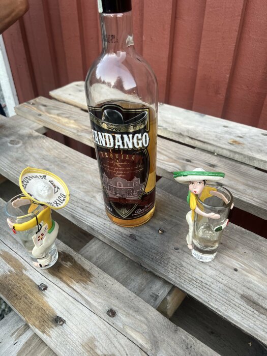 En flaska tequila på en träyta, omgiven av två konstnärligt utformade shotglas.