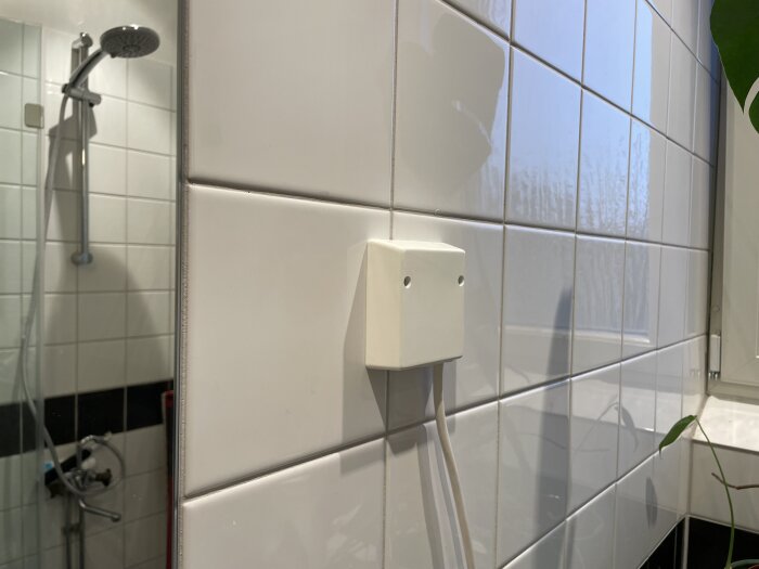 Vit eluttag/dosa installerad mitt i kakelväggen i ett badrum med dusch i bakgrunden, vit vägg med svart bård.