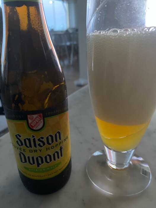 En flaska Saison Dupont öl och ett halvfullt glas med skum på en marmorbänk, bakgrunden visar suddiga konturer av ett rum.