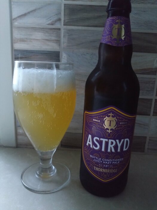 En flaska och ett glas med Astryd Juicy Hazy Pale Ale från Thornbridge på en köksbänk med kakelplattor i bakgrunden.