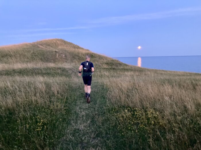 En person joggar på en stig genom en gräsklädd kulle vid havet i skymningen, med en stigande måne reflekterande på vattenytan.