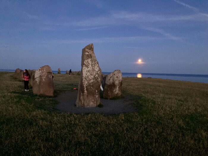 Stenformationer vid kustlinjen i skymningen med fullmåne som reflekteras i havet, människor står intill stenarna.