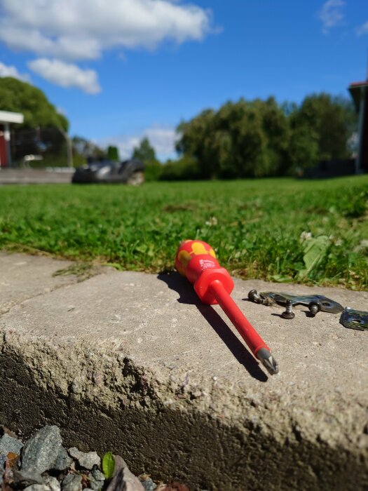 Skruvmejsel, skruvar och metalldelar ligger på en stenplatta i en trädgård, med gräs och träd i bakgrunden under en klarblå himmel.