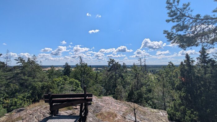 Utsiktsplats med träbänk över en skog och fält under en klarblå himmel med vita moln.