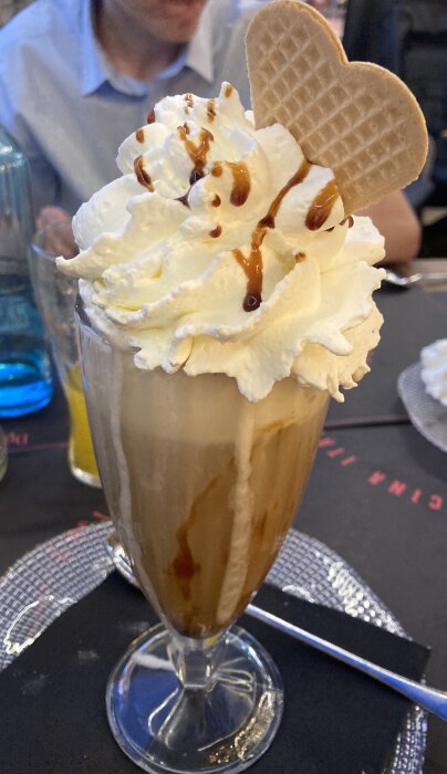 Milkshake med vispgrädde, chokladsås och ett hjärtformat våffelkex, serverad i ett högt glas på ett dukat bord med personer i bakgrunden.