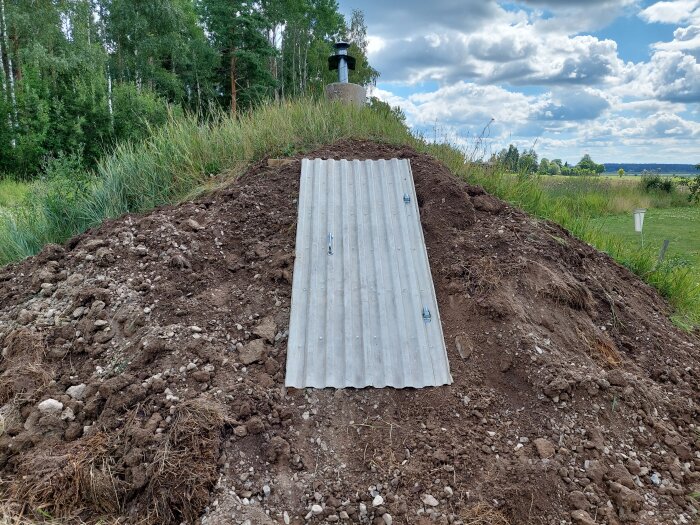 En vit metallskena är placerad i en jordhög som en trappa för att gå ner, omgiven av grön vegetation; trädkant och molnig himmel i bakgrunden.