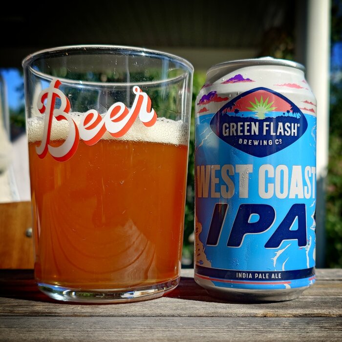 Ett glas med bärnstensfärgad öl bredvid en burk Green Flash Brewing Co. West Coast IPA, fotad utomhus på en solig dag.