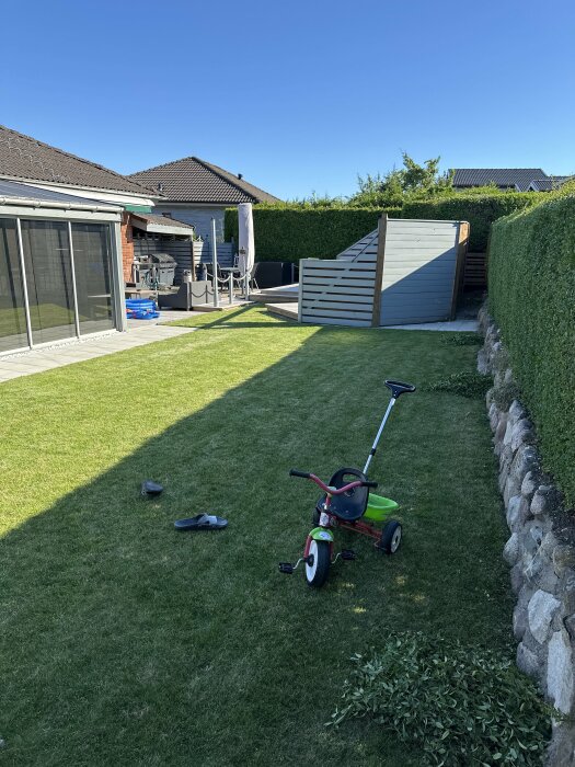 Trädgård med en cykel och tofflor på gräsmattan, ett stenläggningsområde bredvid ett grått plank samt en stenmur och välklippt häck i bakgrunden.