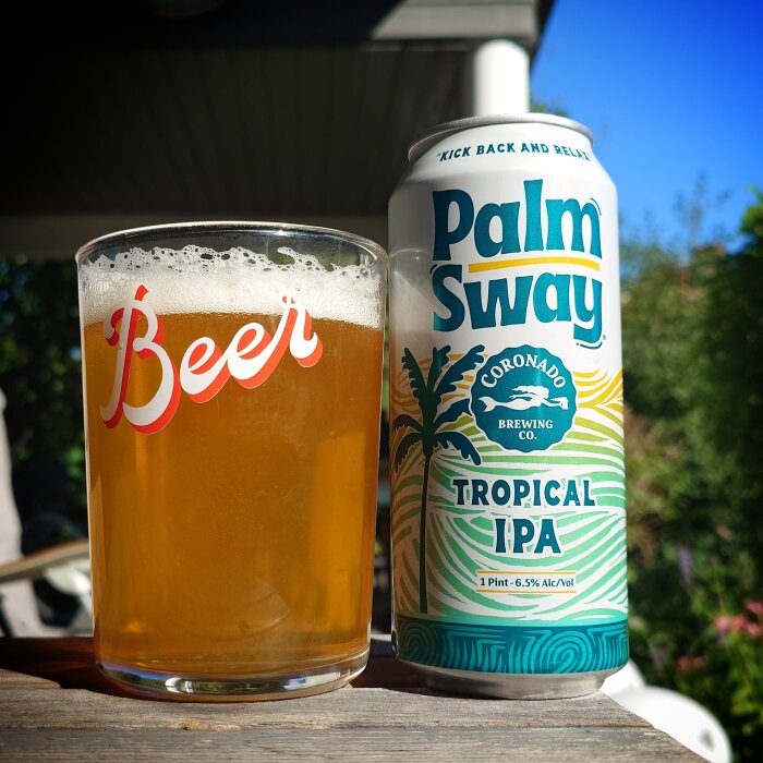 Ölburk med texten "Palm Sway Tropical IPA" och ett glas med öl på ett bord utomhus.