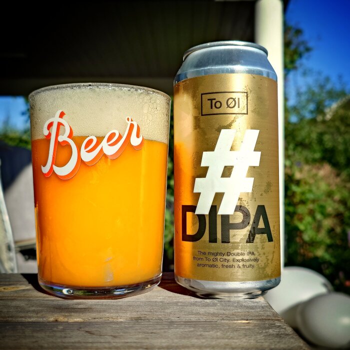 En glas fylld med orange öl med texten "Beer" och en burk To Øl #DIPA i nära en bakgård.