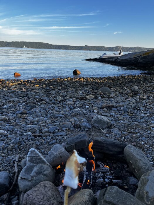 Lägereld på stenig strand vid havet i solnedgång med grillad marshmallow på pinne, båt förtöjd vid klippa i bakgrunden.