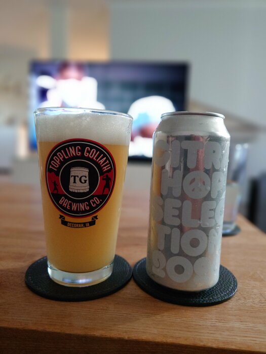 Ett ölglas med logotypen Toppling Goliath Brewing Company och en ölburk med texten "CITRA HOP SELECTION 2020" står på ett bord framför en tänd TV.