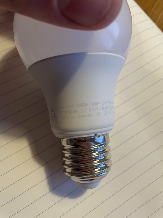 Närbild på en LED-lampa från IKEA som visar specifikationerna: 9W, 806lm, 2700K. Lampan hålls i en hand över ett randigt papper.