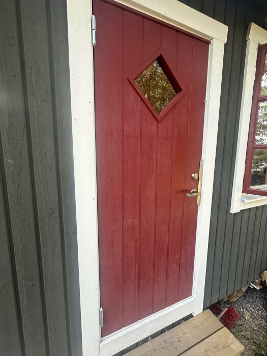 Röd dörr med fyrkantigt fönster mot grå träfasad, vitmålad dörrkarm och solblekt yta, bredvid ett fönster med samma färgsättning.