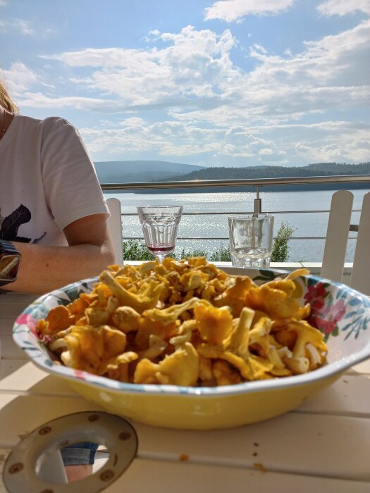 En skål med gula kantareller på ett bord utomhus, med utsikt över en sjö och berg i bakgrunden under en solig dag.