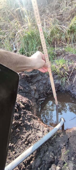 Mark med vattenfylld grop, person håller måttstock som visar cirka 60 cm djup. Våt lerig jord och gräs runt om gropen.
