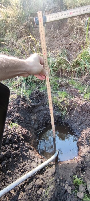 En upprätt mätsticka som hålls i en hand visar ett vattenmätt hål i lerig mark, med gräs och jord i bakgrunden.