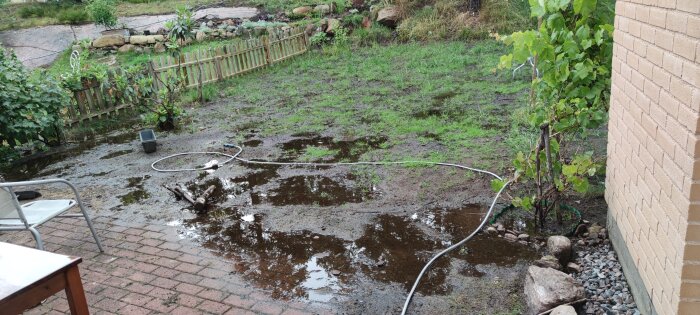 En vattenfylld trädgård med lerig mark vid sidan av ett hus, stängsel i bakgrunden och en slang liggande på marken.