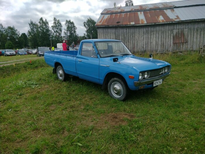 Blå pickup av passlig storlek står parkerad på en grön gräsplan framför en äldre ladugårdsbyggnad med rostiga inslag.