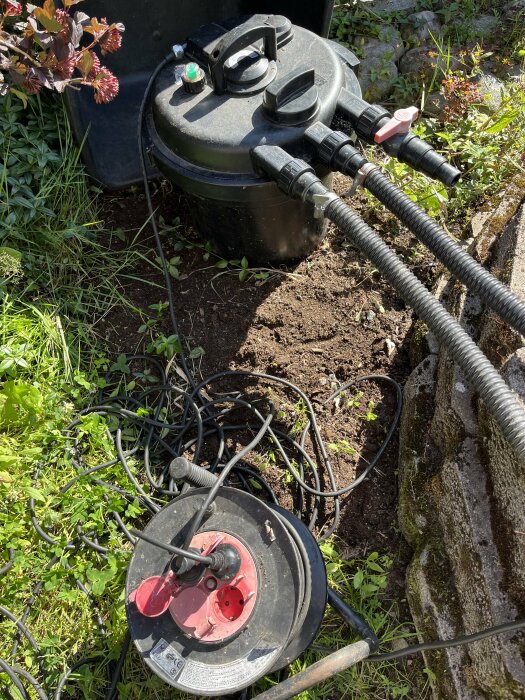 Svart vattentank med kopplade slangar och eldriven pump, bredvid en röd och svart kabelvinda som ligger på marken, omgiven av växtlighet och en stenmur.