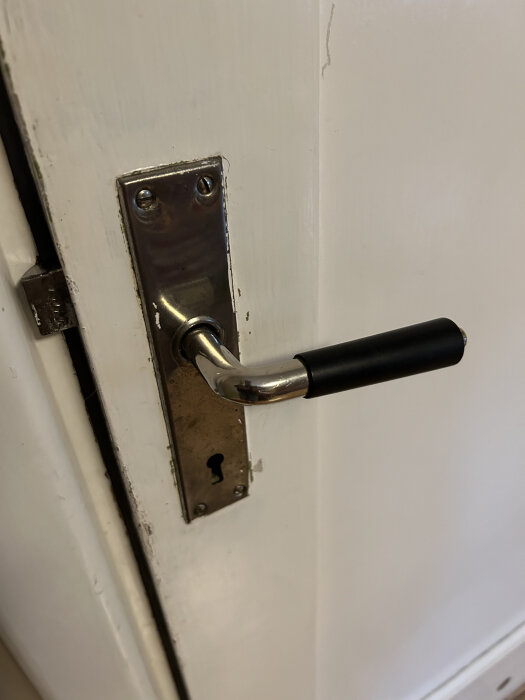Dörrhandtag med svart grepp, eventuellt i bakelit, monterat på en vit dörr med nyckelhål och metallplatta.