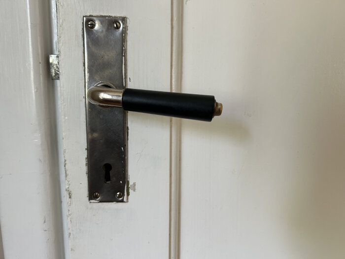 Svart dörrhandtag i metallfäste på en vit dörr, med ett äldre nyckelhål under handtaget.