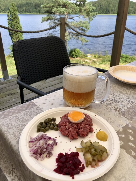 Tartar med rått ägg, kapris, rödlök, rödbetor, ättiksgurka och senap på en papptallrik, tillsammans med en öl på ett bord med utsikt över en sjö.