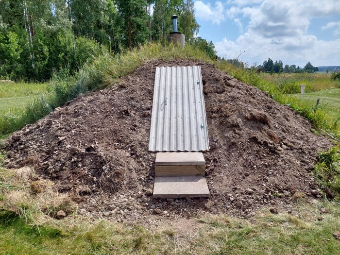 Trappsteg framför en ingång med en plåtdörr på en gräsklädd kulle, omgiven av skog och åkrar under en molnig himmel.