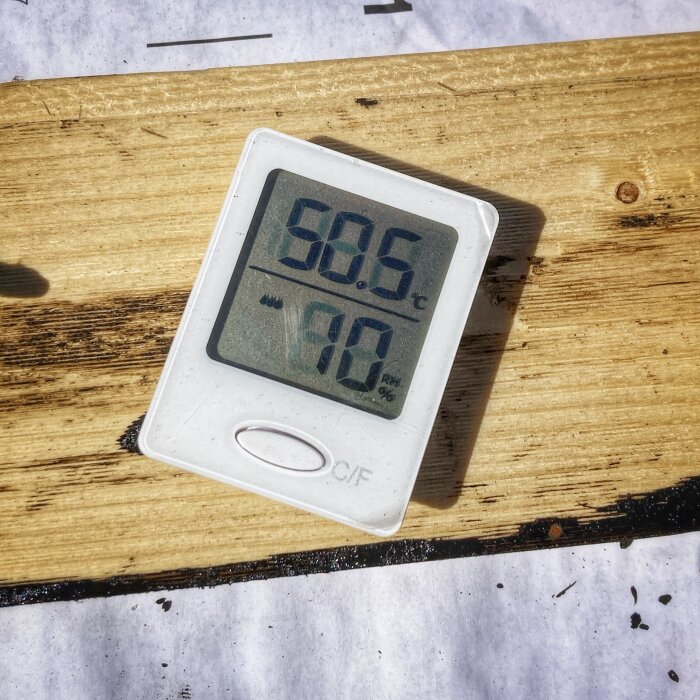 Digital termometer visar 50,5°C och 10% luftfuktighet placerad på en bräda, omgiven av pappmaterial under ett takläggningsprojekt.