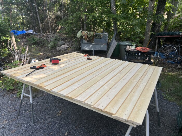 Trädörrar under konstruktion på bockar utomhus med ett fältverktyg och tvingar placerade ovanpå, omgiven av skog och arbetsmaterial.