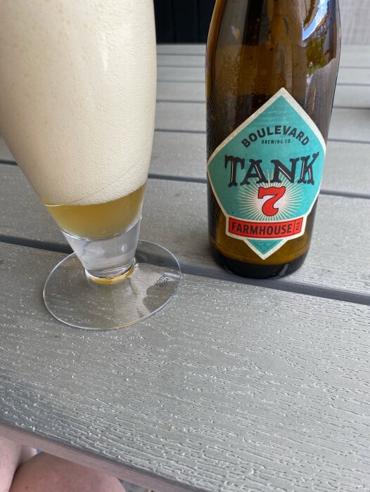 Flaska med Boulevard Brewing Co. Tank 7 Farmhouse Ale och ett upphällt glas med samma öl, med rikligt skum, på ett grått bord utomhus.