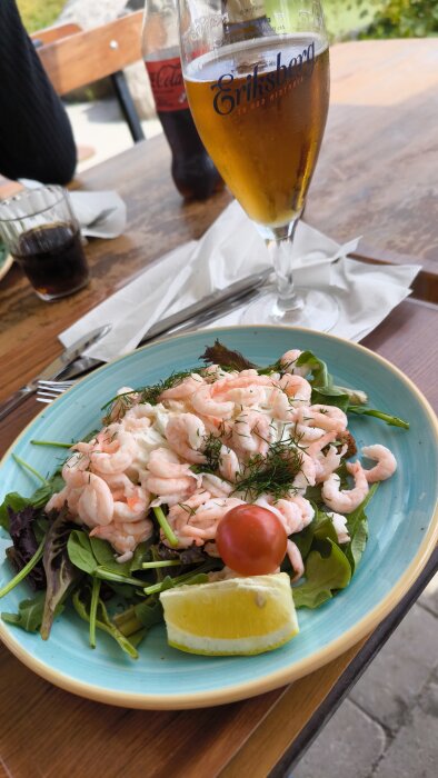 Räksmörgås på en tallrik med sallad och en citronbit, serverad på ett bord tillsammans med ett glas öl och en flaska cola i bakgrunden.