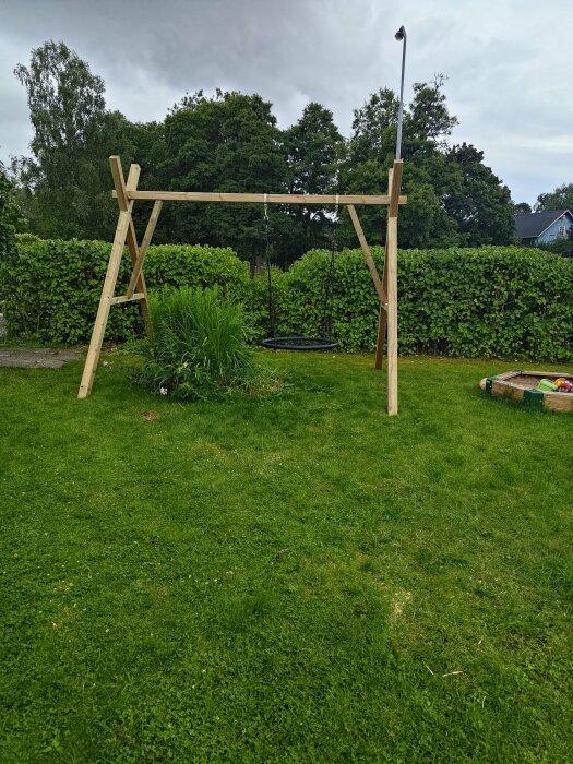 Trägunga på gräsmatta med buskar i bakgrunden. På marken syns blommor som inte har tagits bort än. Treskikts sandlåda synlig till höger.