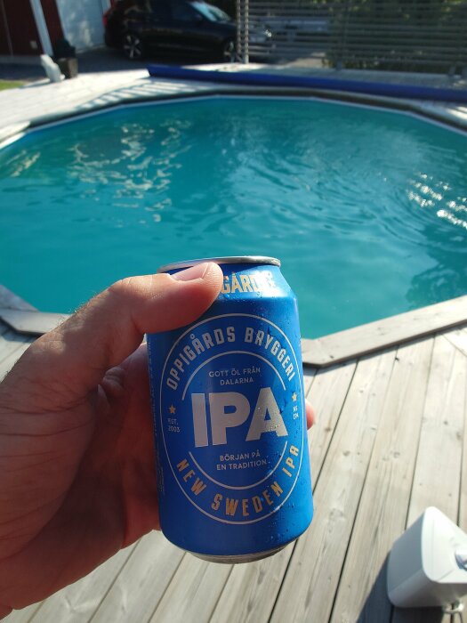 En hand som håller en burk Oppigårds Bryggeri IPA framför en pool med trädäck runt.