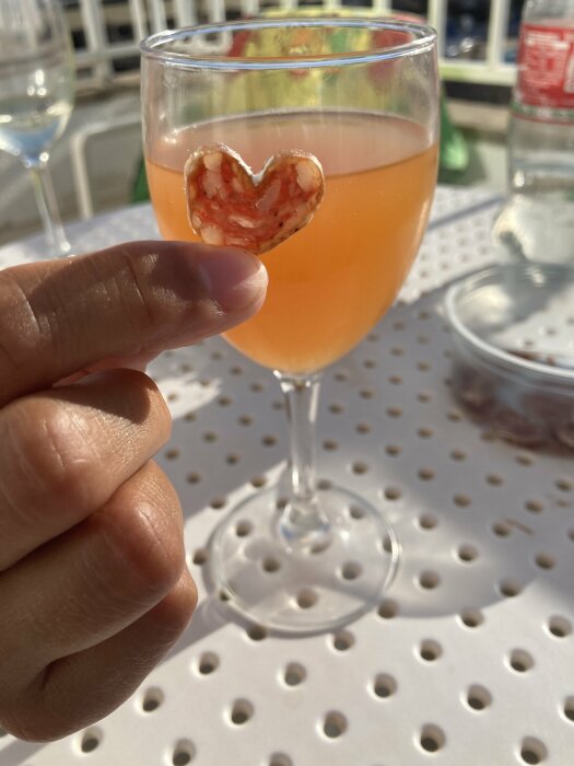 Ett glas med orange dryck på ett perforerat bord, en hand håller en hjärtformad bit av vad som ser ut som korv framför glaset.