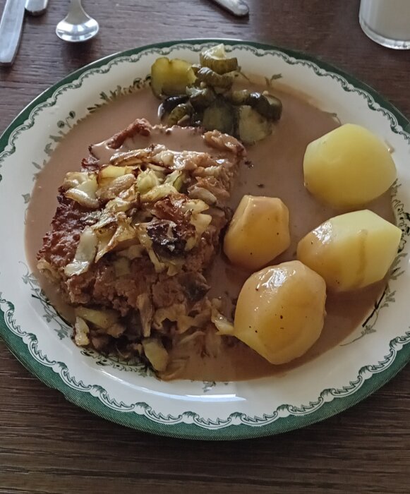 En tallrik med kålpudding, kokt potatis, inlagd gurka och brunsås.