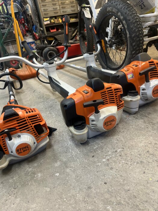 Tre orange/redskap från STIHL ligger på golvet bredvid en cykel i ett välutrustat garage med verktyg och cykelutrustning i bakgrunden.