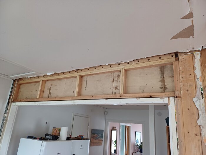 Vägg i kök med en bred dörröppning, där övre delen av väggen rivits för att avslöja 2x4-reglar och plywood utan en synlig balk; takskador syns.