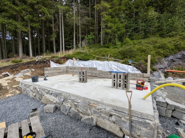 Byggplats med gjuten betongplatta, delvis färdigställd stödmur av leca-block, verktyg och material, omgiven av skog och grus.