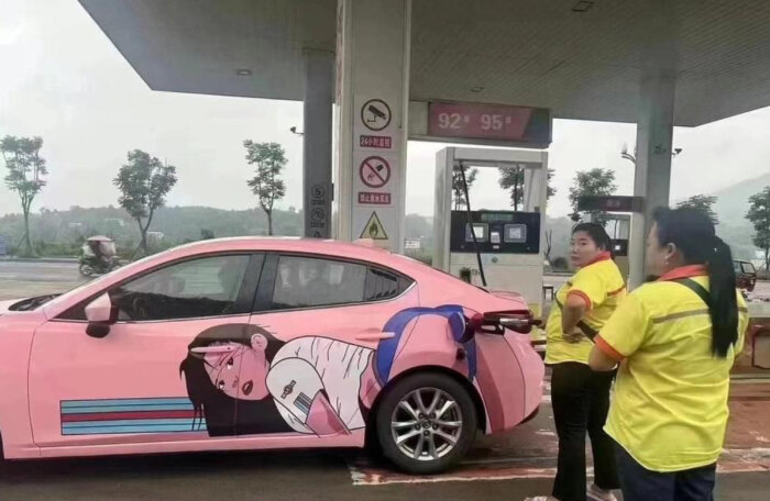 En rosa bil med en animebild på en flicka blev tankad på en bensinmack, medan två personer i gula tröjor står bredvid och tittar på.