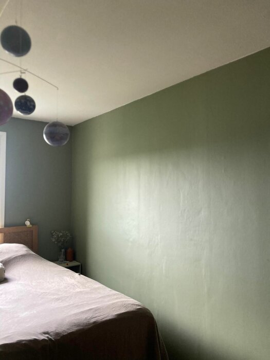 En grön vägg i ett sovrum som visar en del ojämnheter i ytan. I hörnet står en säng och en byrå med några dekorationer.
