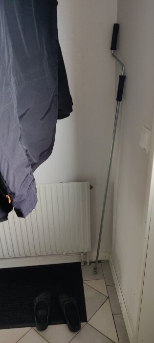 Radiator under ett klädesplagg, två tofflor på en mörk matta och ett verktyg lutat mot väggen på ett klinkergolv i hallen.