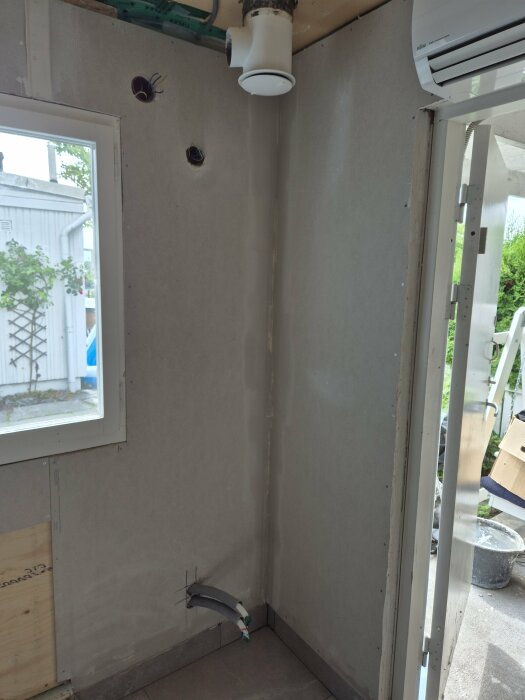 Ett hörn av ett rum med två ventilationsrör och kablar i väggen samt ett fönster och en dörröppning. Ett luftvärmepumpsaggregat är installerat ovanför dörren.