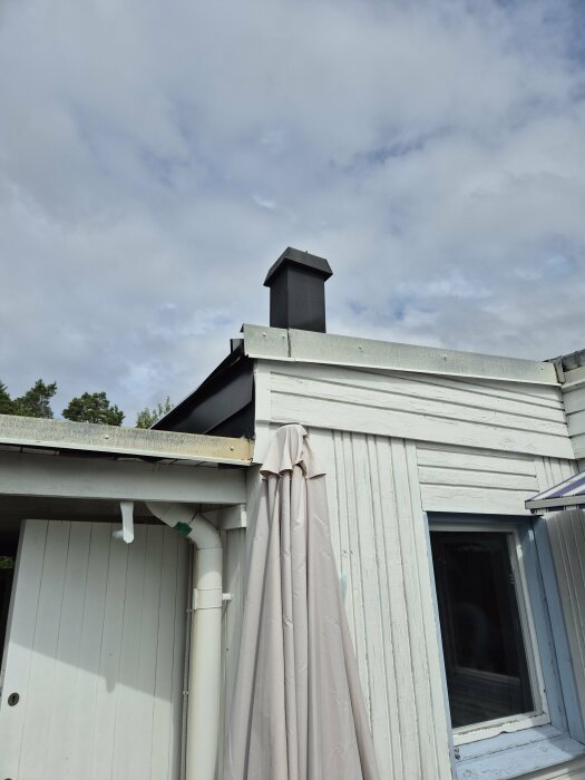 Huset exteriör, vit träfasad med utomhusventilation ovanför ett vitt dagvattenrör, grått plåttak och en stängd dörr till vänster.