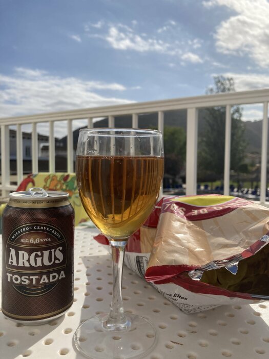 Burk med Argus Tostada öl bredvid ett fyllt vinglas och en öppen chipspåse utomhus på ett bord. Solig dag med blå himmel och några moln i bakgrunden.