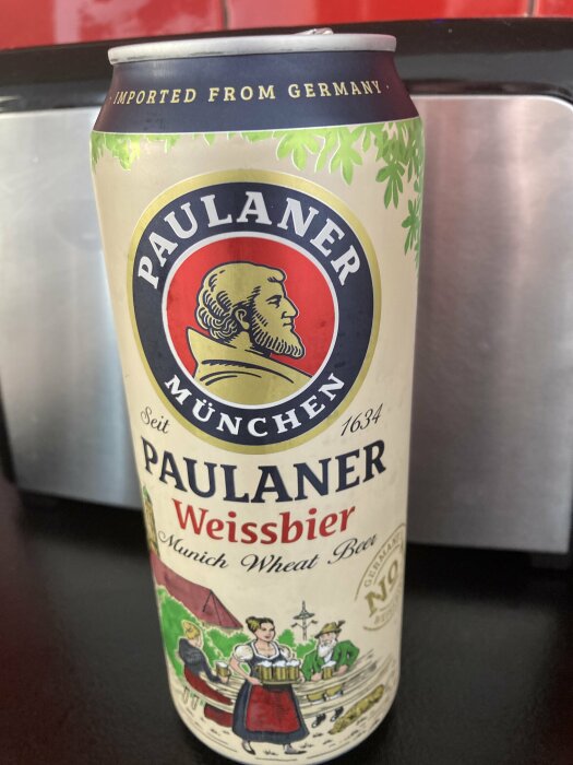 Burk med Paulaner Weissbier, en munich wheat beer, dekorerad med bild av personer i traditionella kläder som serverar öl, texten "imported from Germany" synlig högst upp.