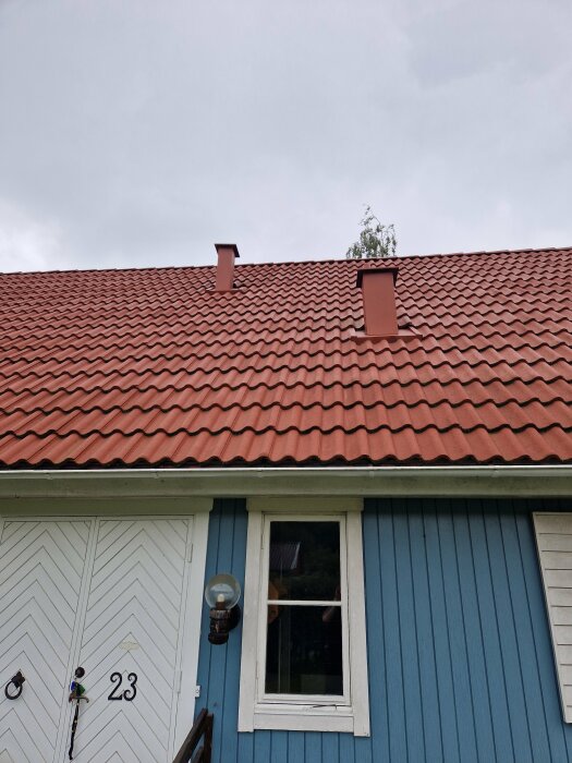 Ett blått 1,5-planshus med rött tak med två självdragskanaler på taket. Huset har en vit dörr med nummer 23 och en fönster bredvid.